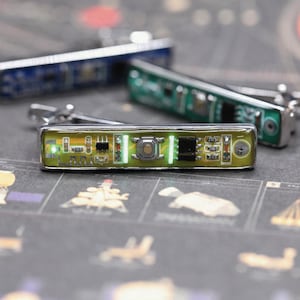 Puede incluir: Un pasador de corbata rectangular que presenta una placa de circuito amarilla con componentes electrónicos visibles, incluyendo un botón central, chips integrados y pequeñas luces verdes brillantes. Un marco metálico rodea el conjunto.