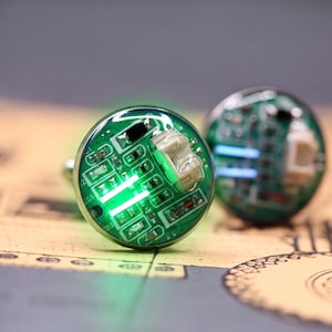 May include: A pair of round cufflinks featuring a green printed circuit board design under a clear dome. The front cufflink displays a bright green LED light illuminating the electronic components, including resistors and a small white connector.