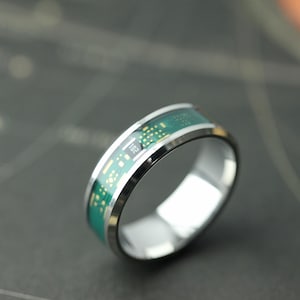 Puede incluir: Un anillo plateado con un diseño de placa de circuito verde. El anillo tiene un acabado pulido y una banda verde con detalles amarillos. El número "102" está impreso en la banda verde. Una joya única.