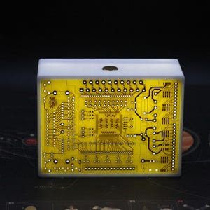 Puede incluir: Una caja de luz rectangular con un diseño de placa de circuito impreso amarilla en el panel frontal. La placa muestra intrincados trazos electrónicos negros, varios puntos de conexión y un patrón central de circuito integrado. Un botón circular está centrado en el borde superior del marco blanco.