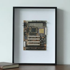 May include: A framed vintage computer motherboard. The circuit board is brown and green with various chips, connectors, and slots. The frame is black and the background is white. A book is visible in the lower left corner.