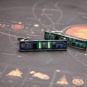 May include: A pair of rectangular cufflinks featuring exposed blue and green printed circuit boards with silver-toned metal frames. Each cufflink displays electronic components, including a central button and two small glowing green LED lights.