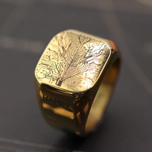 Puede incluir: Anillo dorado con una cara cuadrada con un diseño de placa de circuito en negro. La palabra "HAPPY" está grabada en el lateral. El anillo tiene un acabado pulido y una banda gruesa.