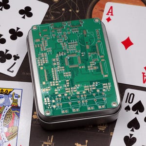 May include: A rectangular metal tin featuring a green printed circuit board design on its lid. The surface displays intricate gold-colored electronic traces, various microchip footprints, and circular mounting holes at each of the four corners.