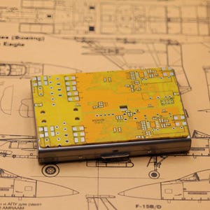 Può includere: Un porta carte rettangolare con un design a circuito stampato giallo. Il porta carte ha una cornice in metallo argentato e poggia su uno sfondo di progetto. Il design del circuito stampato presenta vari componenti e percorsi elettronici.