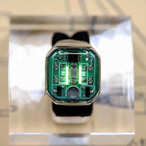 May include: A ring featuring a rectangular green circuit board face with exposed electronic components, including a central silver crystal oscillator labeled 26.000 MHz, two vertical light strips, and a small circular speaker component.