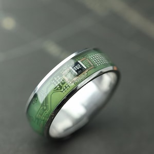 May include: A silver ring with a green circuit board design. The ring has a clear resin overlay, displaying the intricate electronic components. The ring has the number "102" printed on it.