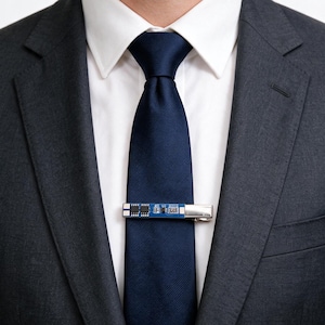 May include: A dark navy blue necktie worn with a white collared shirt and a charcoal gray suit jacket. A silver-toned tie clip featuring a small blue rectangular circuit board component with visible black microchips is attached to the necktie.