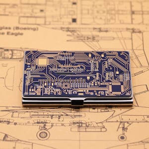 以下が含まれることがあります： 金色の回路基板デザインが施された、長方形のネイビーブルーのカードケース。 カードケースはシルバーの金属フレームで、前面には「BOHIMU」の文字がプリントされています。 背景はベージュの設計図です。