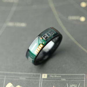 Puede incluir: Un anillo de banda negra con una incrustación de placa de circuito verde, decorado con componentes electrónicos plateados y dorados, incluyendo un pequeño chip rectangular marcado con el número 102, sobre una superficie oscura y pulida.