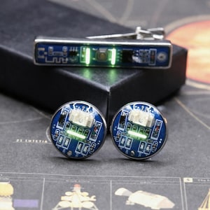 May include: A rectangular tie bar and two circular cufflinks featuring blue printed circuit board patterns with integrated green LED lights. The electronic components are mounted on silver-toned metal bases, showcasing a technical design.