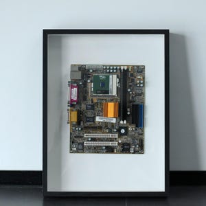 May include: A framed computer motherboard featuring a central Intel processor, a bright orange heat sink, various integrated circuits, and multiple expansion slots. The circuit board is mounted against a white background within a black frame.