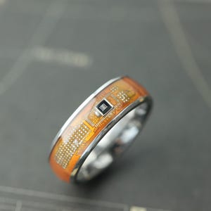 May include: A silver ring with an orange circuit board design. The ring features a rectangular black component with the number "1001" printed on it. The circuit board design is visible on the ring's surface.