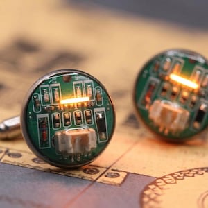May include: A pair of circular cufflinks featuring green printed circuit boards with visible electronic components, including small resistors and a white connector, each illuminated by a single horizontal orange light bar.