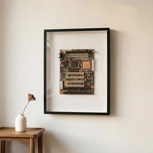 May include: A framed computer motherboard, displaying intricate electronic components. The board is enclosed in a black frame, hung on a white wall. A small vase with a dried flower is on a wooden table beneath the artwork.
