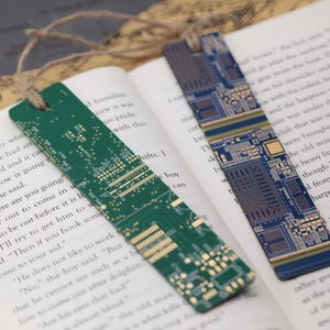 May include: Two rectangular bookmarks with a circuit board design. One is green, the other is blue. Both have a hole at the top with a natural twine string. The bookmarks are resting on the pages of an open book.