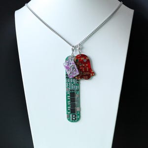 May include: A silver chain necklace featuring three charms made from recycled circuit boards. The charms include a long green rectangular board, a small purple rectangular component, and a red ghost-shaped piece with gold circuitry patterns.