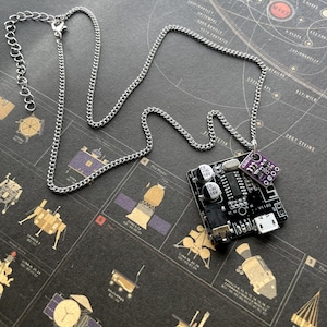 May include: A silver chain necklace with a pendant featuring a black circuit board and a small purple component. The necklace is displayed on a dark background with illustrations of spacecraft and celestial bodies.