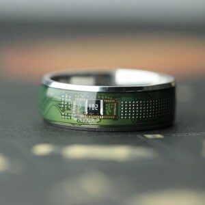 May include: A silver ring with a green circuit board design. The ring features a small rectangular component with the number "102" printed on it. The ring has a polished silver band and a dark background.