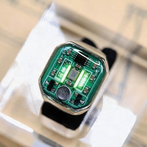 Könnte beinhalten: Ein silberfarbener Ring mit einer rechteckigen Fassung, die eine grüne Leiterplatte zeigt. Das elektronische Bauteil enthält einen zentralen Metallchip mit der Aufschrift 26.000 MHZ, kleine Widerstände und zwei leuchtende Streifen.
