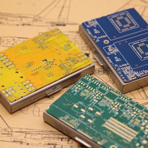 Puede incluir: Tres carteras metálicas con diseños de circuitos impresos en amarillo, azul y verde. Las carteras tienen un marco plateado y están dispuestas sobre un fondo de planos arquitectónicos. Los patrones de las placas de circuito son intrincados y detallados.