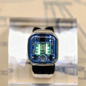 Pode incluir: Um anel de tom prateado com uma face retangular que apresenta uma incrustação de placa de circuito impresso azul. O centro da placa exibe componentes eletrônicos, incluindo um pequeno oscilador de cristal prateado marcado 25.000 MHZ e duas luzes verdes verticais.