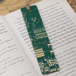 Op de afbeelding: Groen printplaat bladwijzer met een jute touwtje. De rechthoekige bladwijzer toont een gedetailleerd elektronisch circuitontwerp met goudkleurige componenten. Het ligt in een boek met gedrukte tekst.