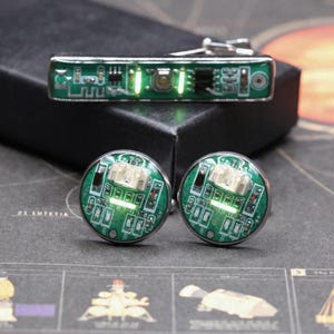 May include: A set of accessories featuring a rectangular tie bar and two circular cufflinks. Each piece is crafted from green printed circuit boards with visible electronic components, metallic silver hardware, and integrated glowing green LED lights.