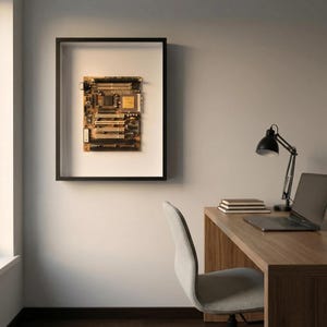 Könnte beinhalten: Eine gerahmte Computer-Hauptplatine mit goldenen und braunen Komponenten hängt an einer weißen Wand. Der schwarze Rahmen umgibt die Leiterplatte und schafft ein modernes, technikbasiertes Kunstwerk. Im Hintergrund befindet sich ein Schreibtisch mit Laptop und Lampe.