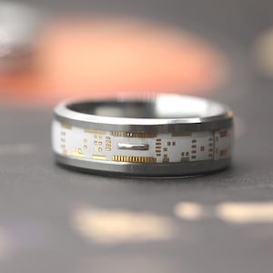 Könnte beinhalten: Ein silberner Ring mit einem weißen Band, das ein goldenes Leiterplattendesign aufweist. Der Ring hat eine polierte Oberfläche und eine moderne, tech-inspirierte Ästhetik. Ein einzigartiges Schmuckstück.