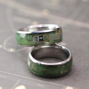 May include: Two silver-toned rings featuring a green circuit board pattern inlay. The top ring displays a small black electronic component with the white text 102 printed on its surface, surrounded by intricate gold-colored circuitry lines.