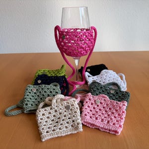 Könnte beinhalten: Ein klares Weinglas in einem leuchtend pinkfarbenen Häkelhalter mit langem Trageriemen. Um das Glas herum liegen weitere Häkelhalter in verschiedenen Farben, darunter Beige, Hellrosa, Dunkelgrün, Weiß, Schwarz und Marineblau.