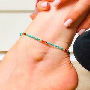 以下が含まれることがあります： 赤と金色のアクセントが付いた、ターコイズ色のビーズアンクレット。 アンクレットは細いコードに通されており、足首に着用されています。繊細なジュエリーです。