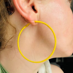 May include: A close-up of a person's ear wearing a large, bright yellow hoop earring. The earring is a simple, circular design, and the vibrant color contrasts with the person's skin tone. The earring is the focal point of the image.