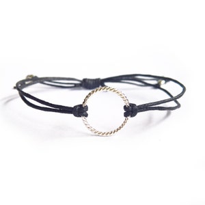 Könnte beinhalten: Ein schwarzes Armband aus Schnur mit einem silbernen Kreisanhänger. Der Kreisanhänger ist gedreht und hat eine glatte Oberfläche.