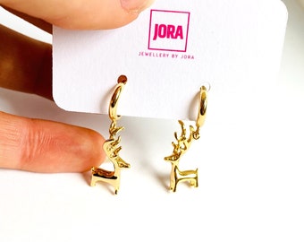 May include: Gold hoop earrings with a small gold reindeer charm hanging from each hoop. The earrings are displayed on a white card with the text "JORA JEWELLERY BY JORA".