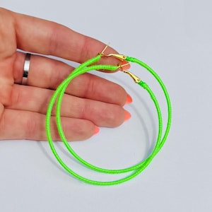 Op de afbeelding: Een paar neon groene oorringen. De hoepels zijn gewikkeld in een felgroene draad en hebben goudkleurige sluitingen. De oorbellen worden getoond tegen een lichte achtergrond.