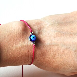 May include: A pink string bracelet featuring a blue and white evil eye charm. The charm is round with a dark blue center and a light blue iris. The bracelet is tied on the wrist with a simple knot.