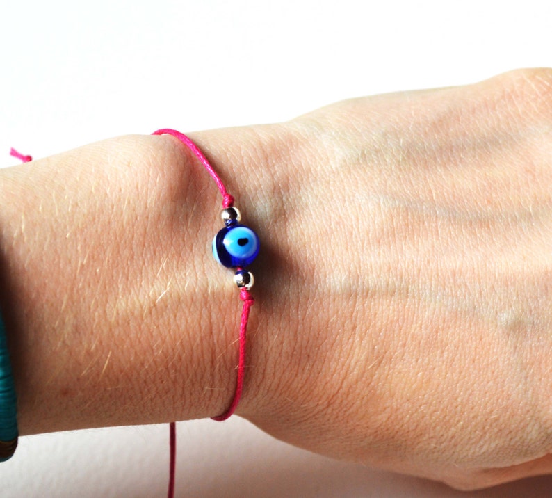 May include: A pink string bracelet featuring a blue and white evil eye charm. The charm is round with a dark blue center and a light blue iris. The bracelet is tied on the wrist with a simple knot.