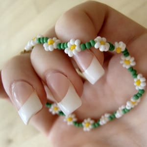 May include: A white and green beaded bracelet with daisy flower charms. The bracelet is on a hand with white nail polish.