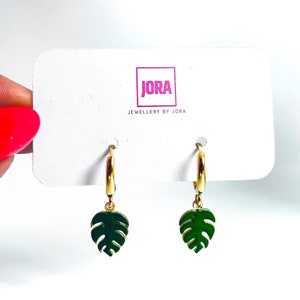 May include: Gold hoop earrings with a green monstera leaf charm. The earrings are on a white card with the text "JORA JEWELLERY BY JORA".