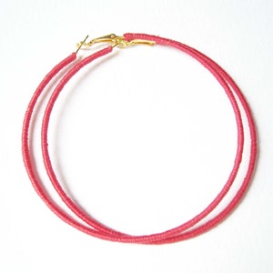 May include: A pair of large hoop earrings made of pink thread. The earrings have gold-toned clasps.