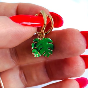 May include: Gold hoop earrings with a green monstera leaf charm. The earrings are shown on a hand with red nail polish.