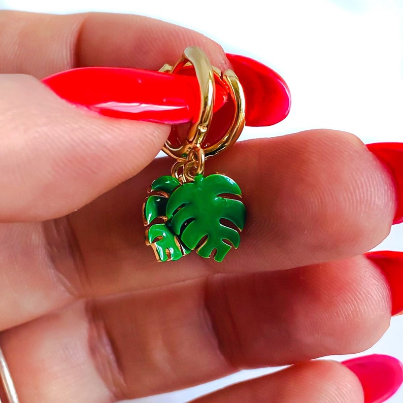 May include: Gold hoop earrings with a green monstera leaf charm. The earrings are shown on a hand with red nail polish.