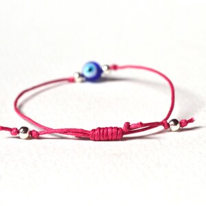 May include: A pink string bracelet featuring a blue and white evil eye bead and silver-colored beads. The bracelet is adjustable with a sliding knot closure. The evil eye bead is a focal point, adding a touch of protection and style.