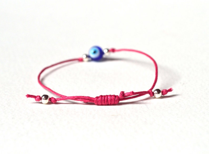 May include: A pink string bracelet featuring a blue and white evil eye bead and silver-colored beads. The bracelet is adjustable with a sliding knot closure. The evil eye bead is a focal point, adding a touch of protection and style.
