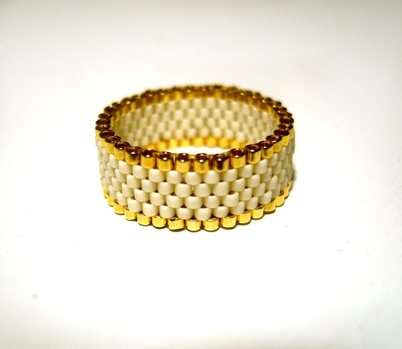 K&ouml;nnte beinhalten: Ein wei&szlig;er Perlenring mit goldener Einfassung. Der Ring besteht aus kleinen, quadratischen Perlen und hat ein schlichtes, elegantes Design.