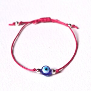 May include: A pink string bracelet featuring a blue and white evil eye charm. The bracelet has adjustable sliding knots and small silver beads. This handmade bracelet is a symbol of protection.
