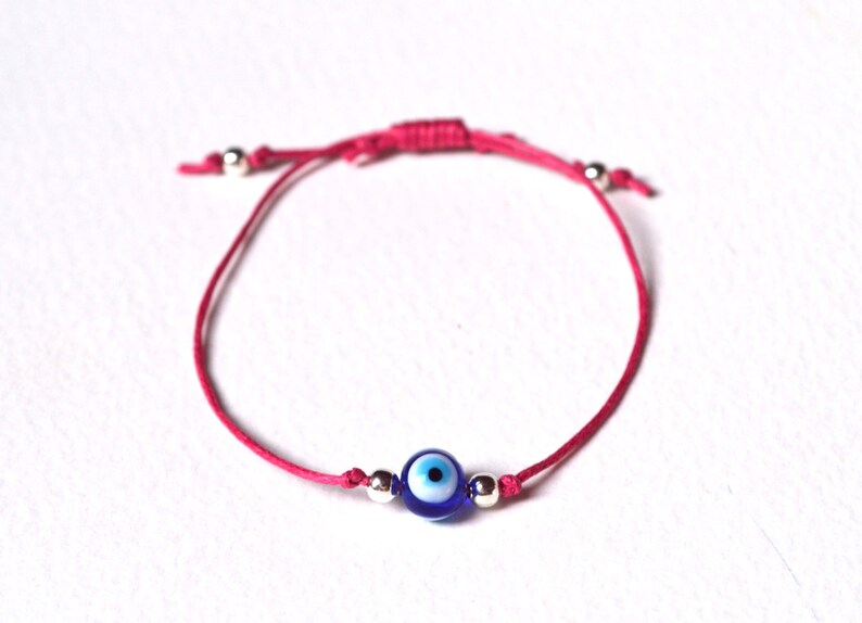 May include: A pink string bracelet featuring a blue and white evil eye charm. The bracelet has adjustable sliding knots and small silver beads. This handmade bracelet is a symbol of protection.