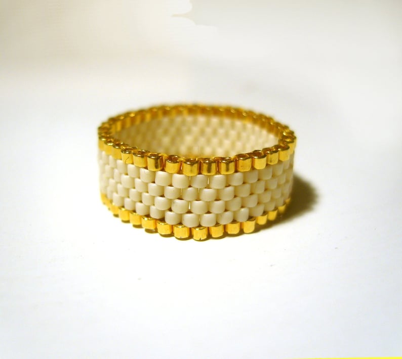 K&ouml;nnte beinhalten: Ein wei&szlig;er Perlenring mit goldenen Akzenten. Der Ring besteht aus kleinen, runden Perlen, die in einem Muster miteinander verwoben sind. Die goldenen Akzente befinden sich an den R&auml;ndern des Rings.